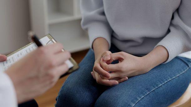 A person in jeans and a sweater sits with hands clasped, facing someone taking notes on a clipboard.