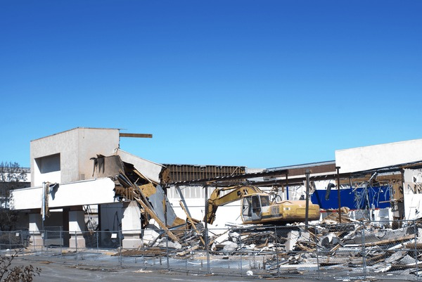 Excavator demolishing a white building against a clear blue sky; debris scattered.