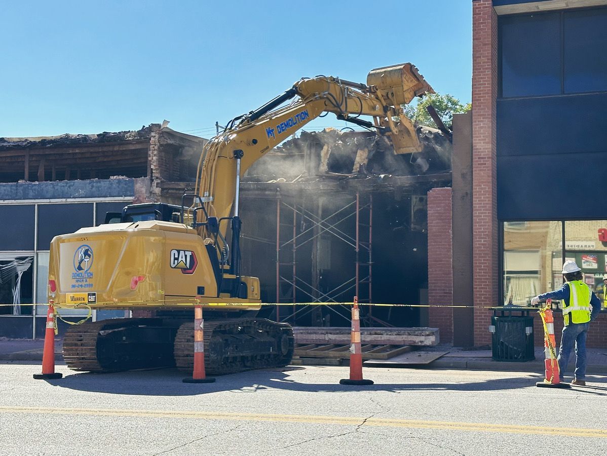 Construction zone with orange barriers and a sign in a dirt area, with buildings in the background.