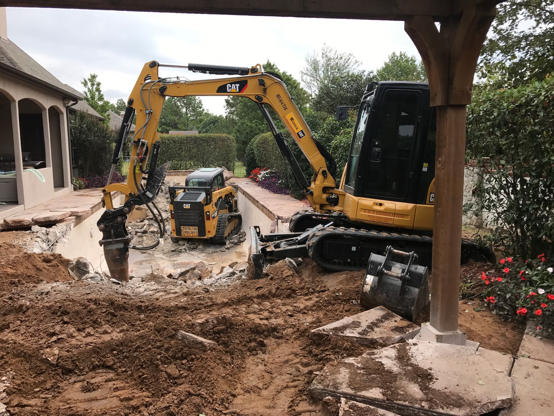 Yellow excavator demolishing a two-story house, debris scattered, blue sky background.