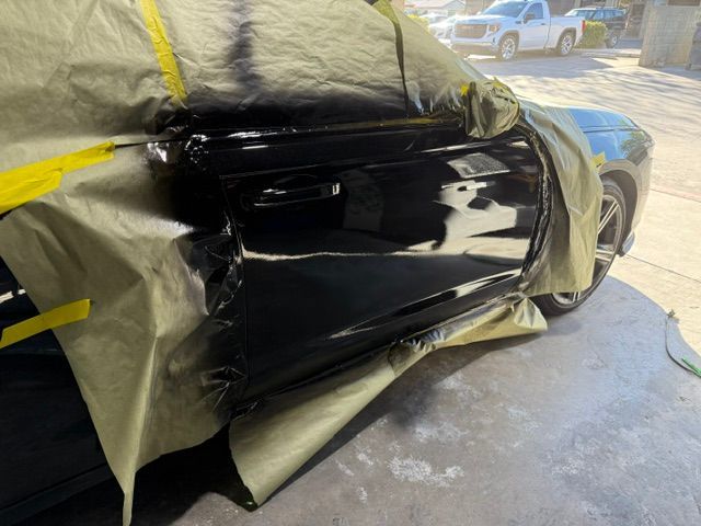Black car door being painted, taped off with yellow tape and covered with plastic.