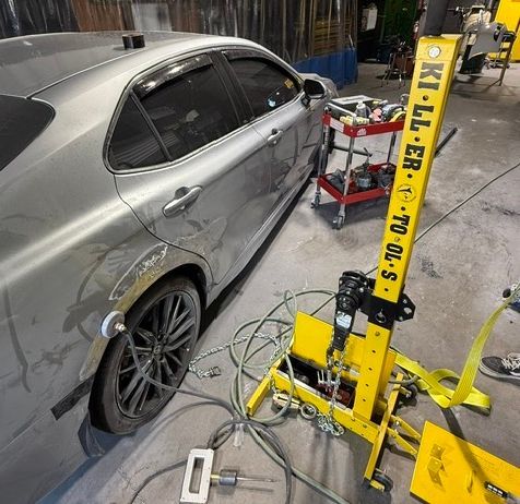 Silver car being repaired by a yellow dent puller in a workshop.