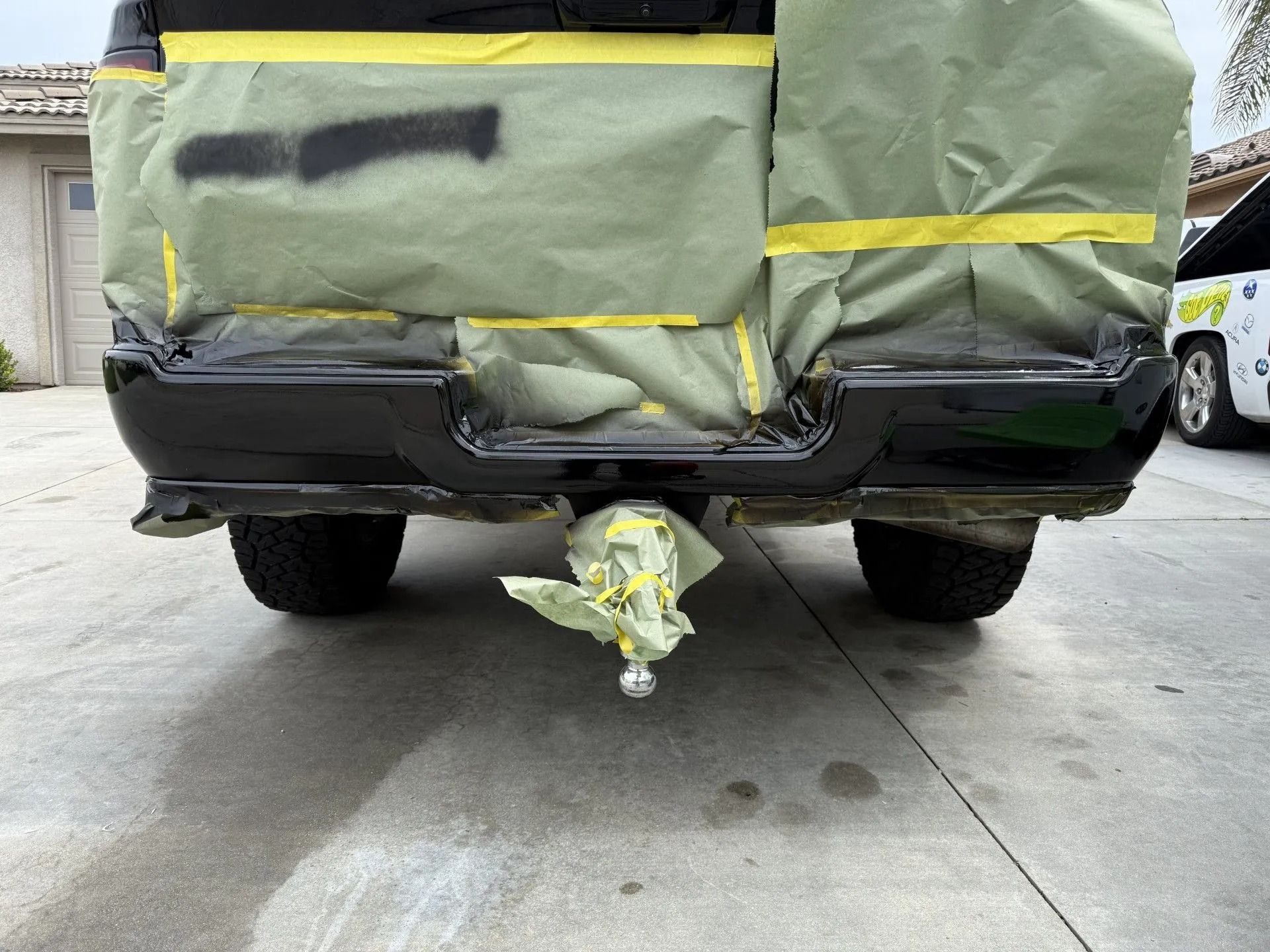 A dark gray Tesla being prepped for paint, with sanded areas visible on its side, parked in a driveway under a canopy.
