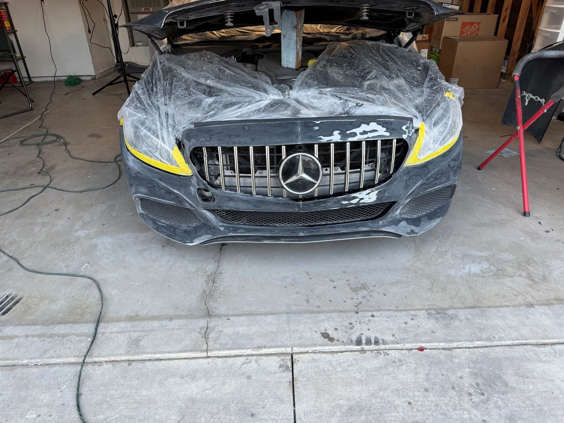 A black Mercedes with the front end covered in plastic and taped, likely undergoing repair in a garage.
