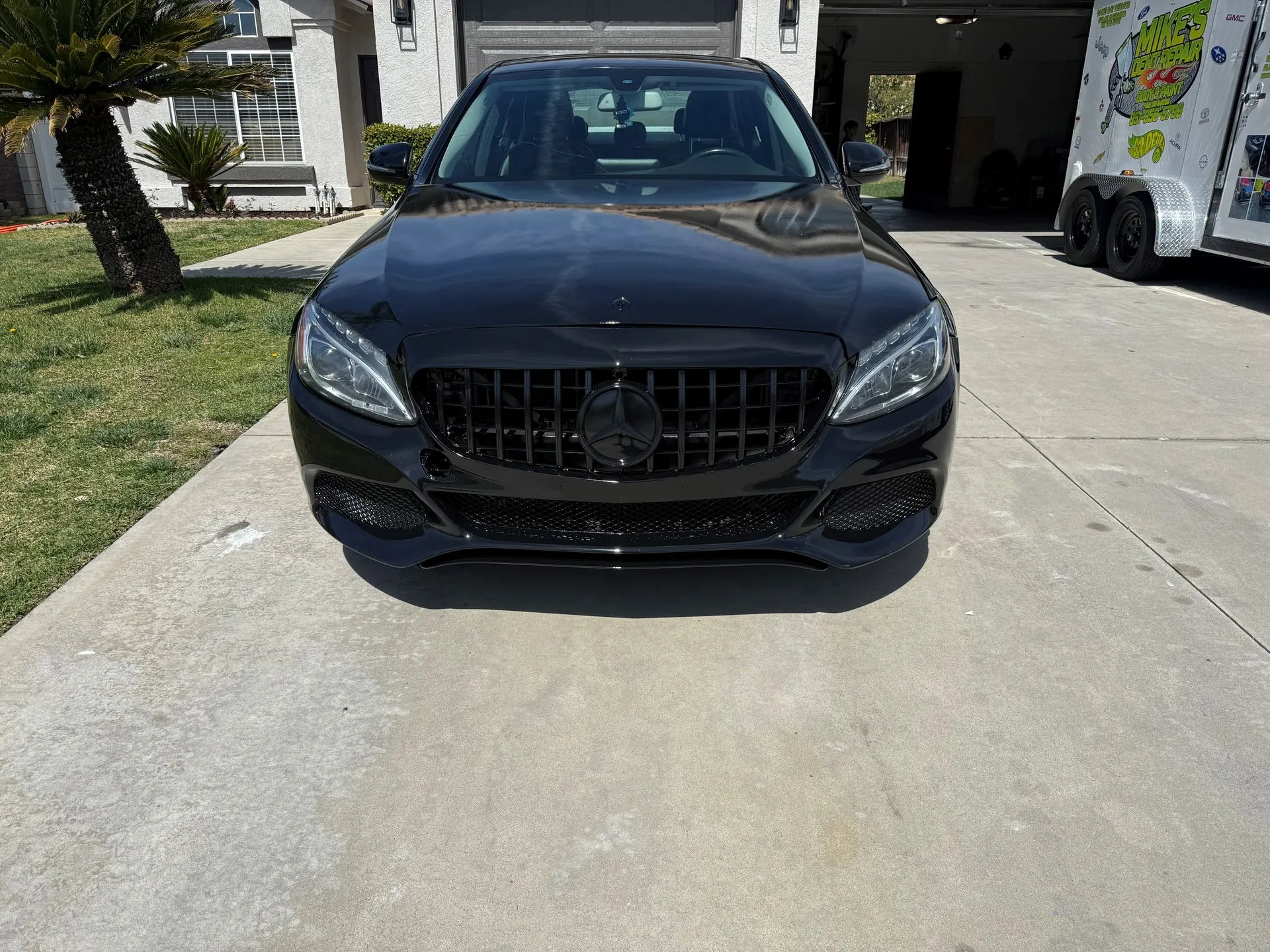 Black Mercedes-Benz sedan parked on a driveway, facing forward. The car has a black grill and is in front of a house with a trailer to the right.