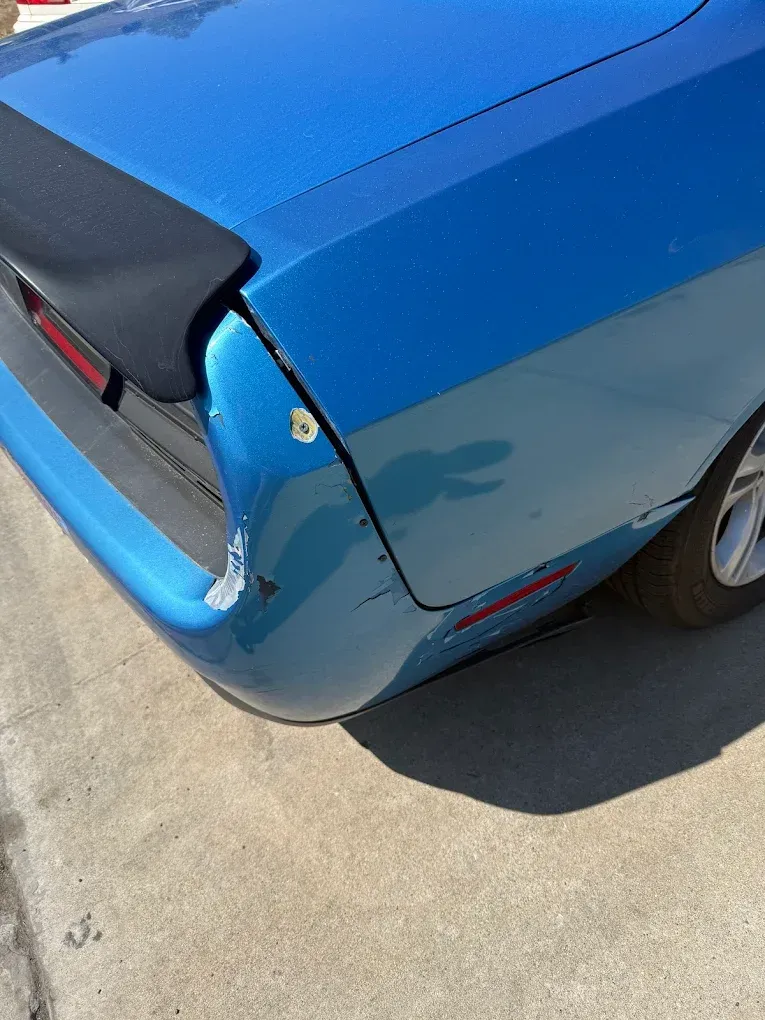Blue car's rear corner with damage to the bumper and body panel. A black spoiler is visible above.