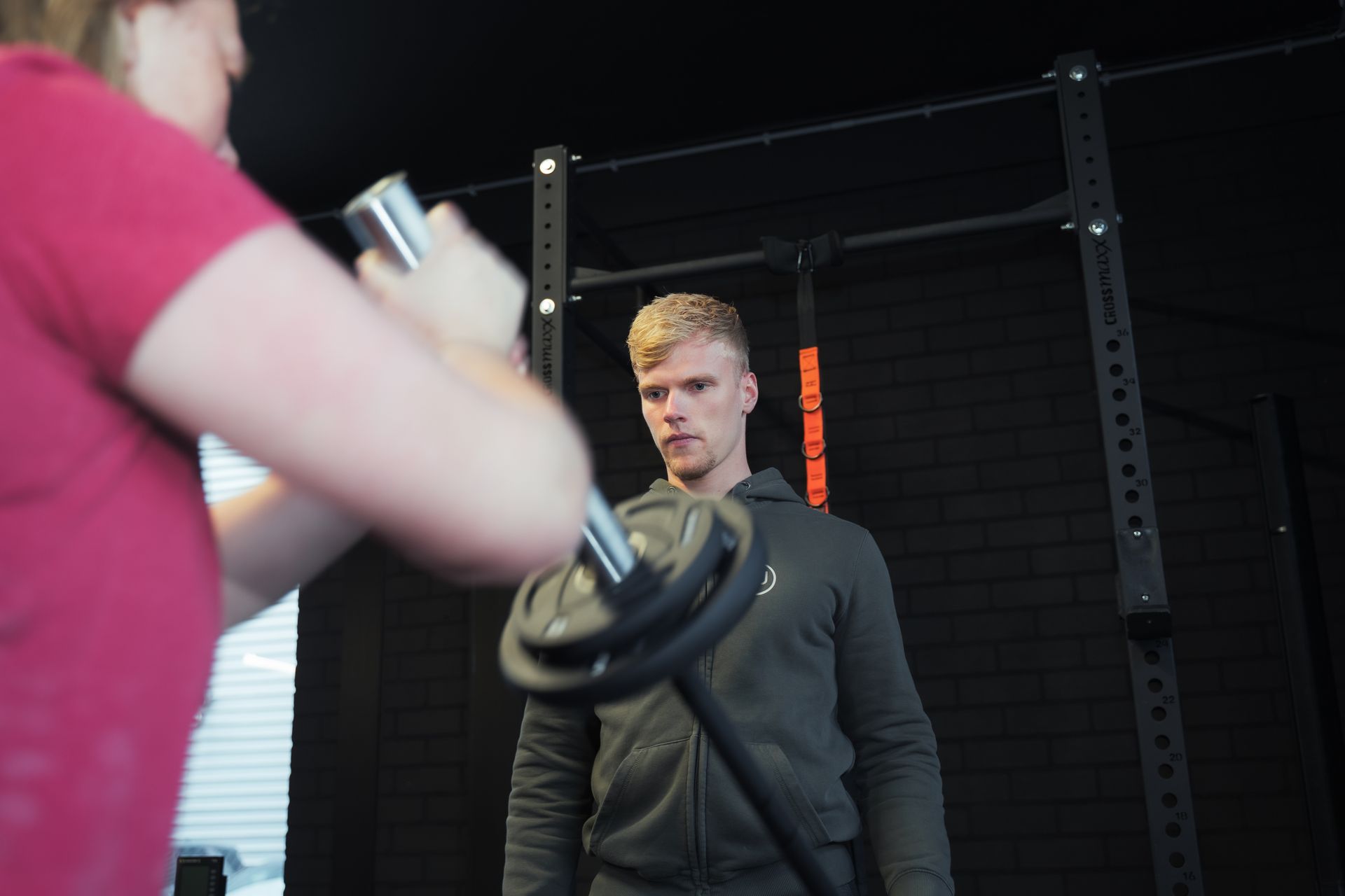 Een man kijkt toe hoe iemand in een roze shirt een halter met gewichtsschijven vasthoudt in een sportschool.