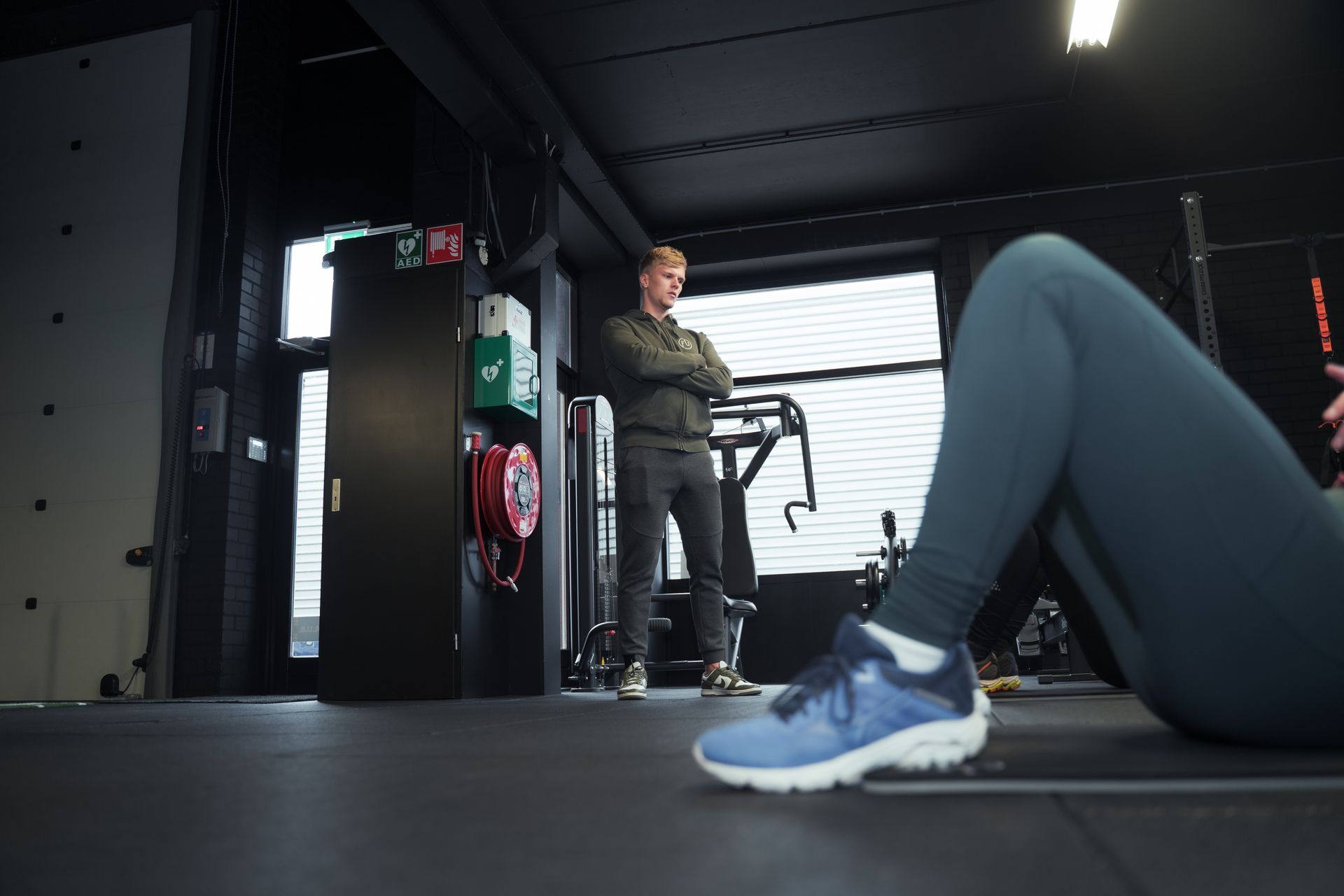 Een man in de sportschool kijkt naar iemand die aan het trainen is. De man heeft zijn armen over elk