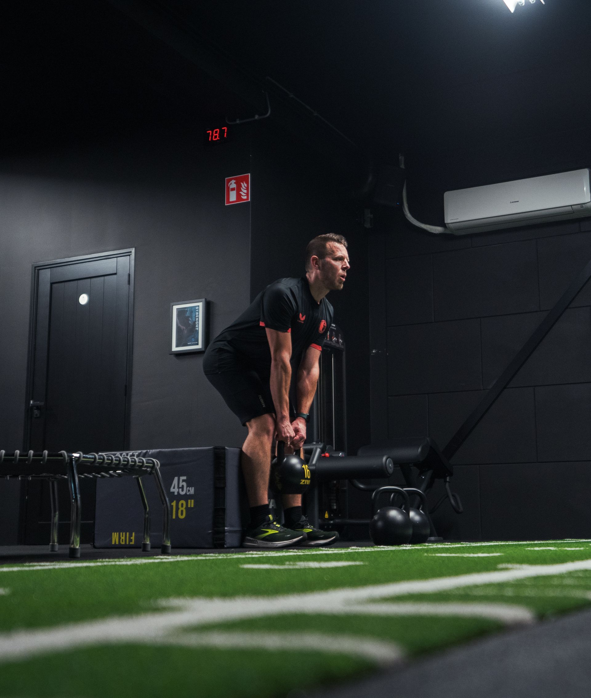 Man in sportschool tilt kettlebell op, zwarte ondergrond, groene grasmat.