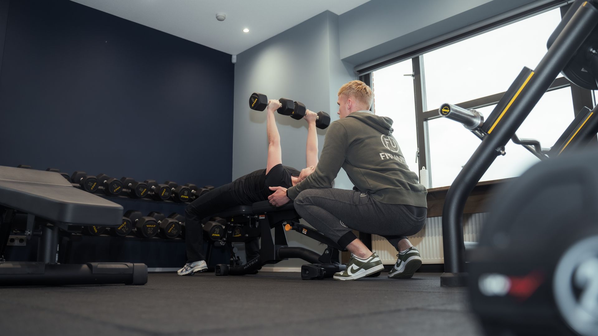 Een persoon tilt een halter op een bankje, geholpen door een trainer, in een sportschool.