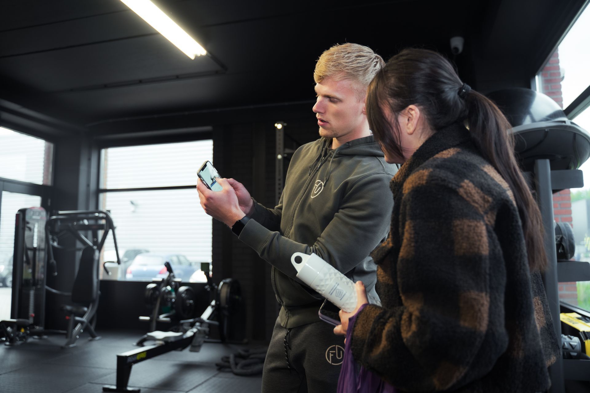 Blonde man in sportkleding laat zijn telefoon zien aan een vrouw in een geruite jas; ze zijn in een sportschool met apparatuur.
