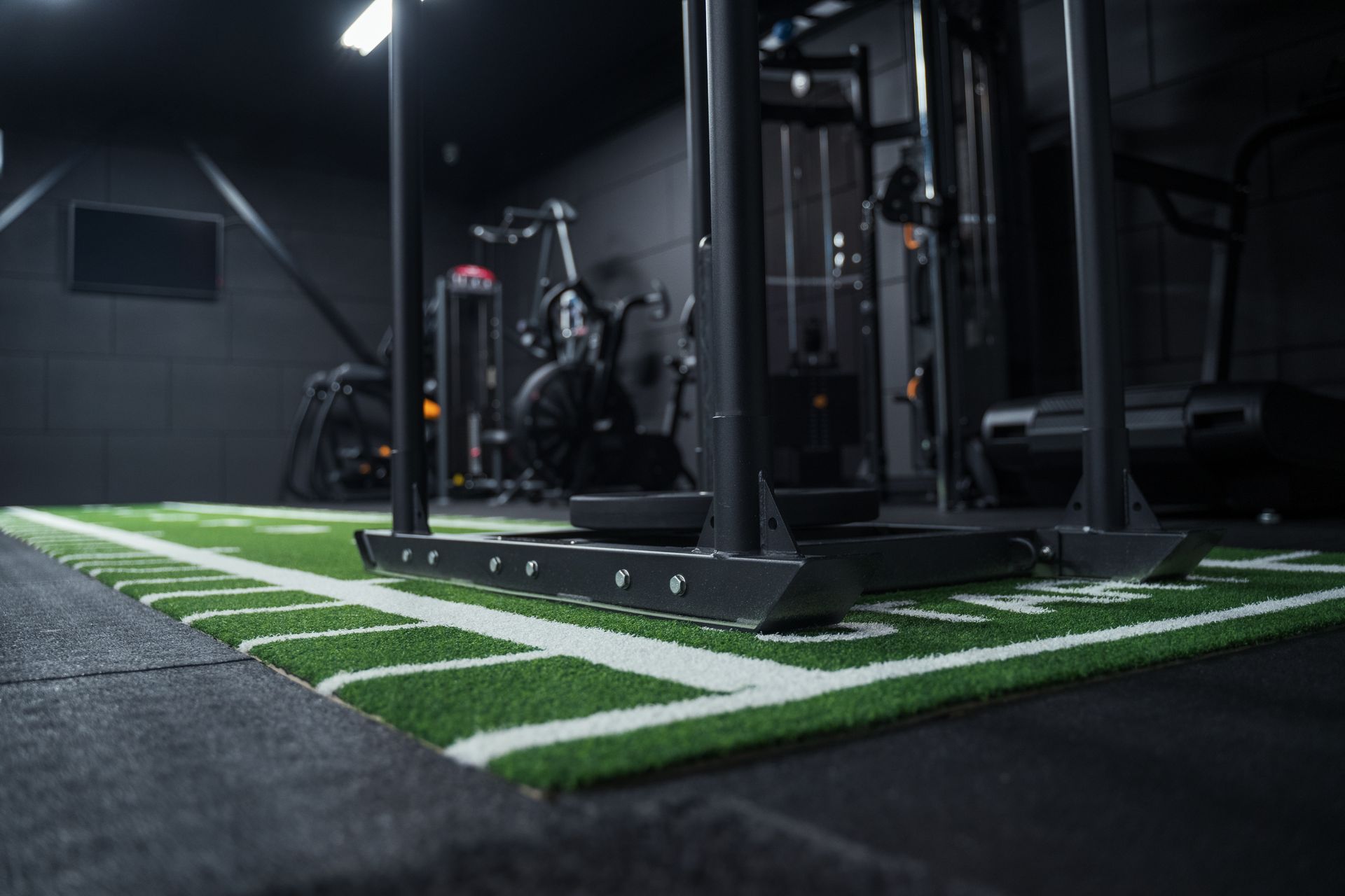 Sportzaalinterieur met zwarte slee op kunstgrasveld met yardlijnen. Op de achtergrond staat diverse fitnessapparatuur.