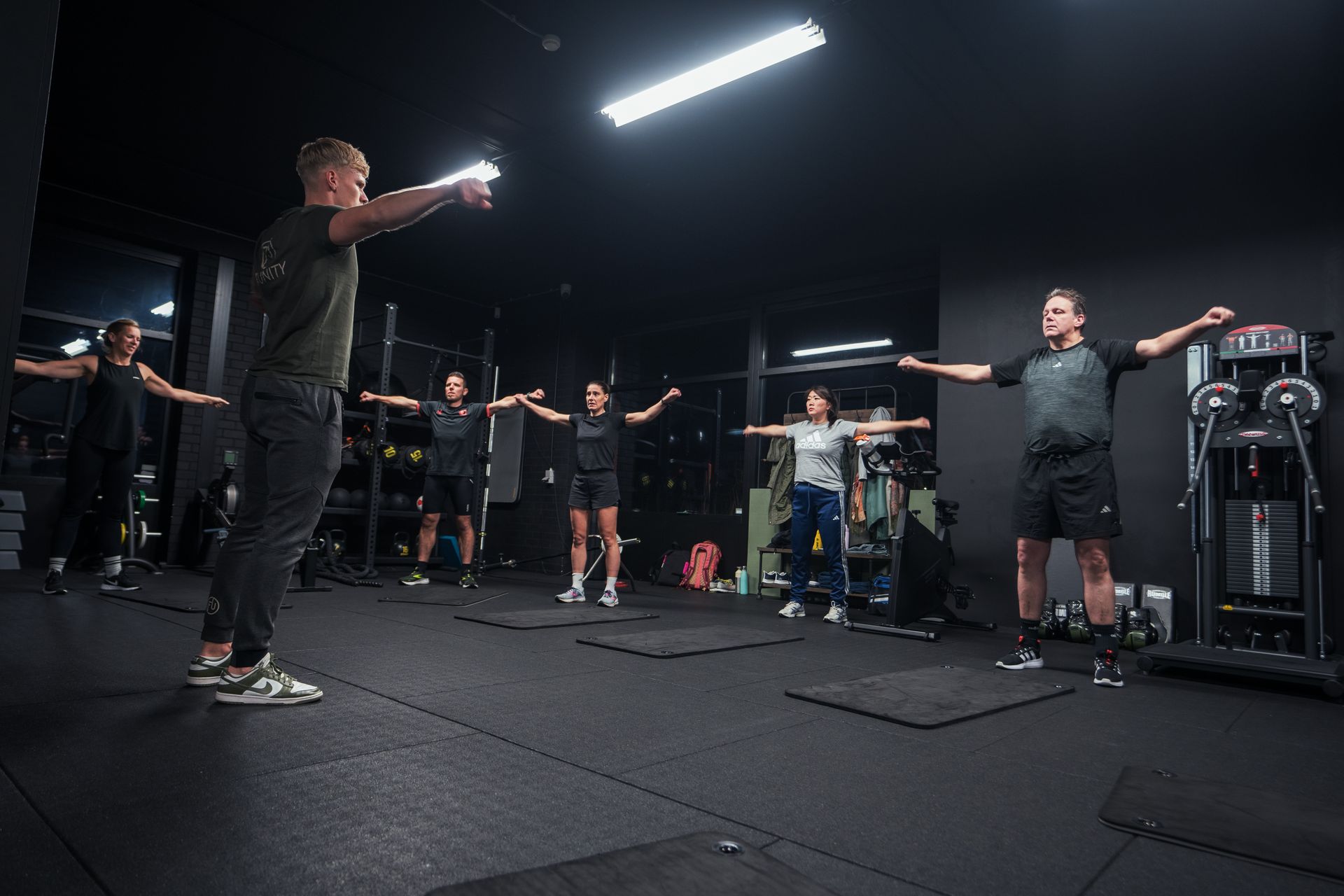 Mensen in een sportschool met uitgestrekte armen, onder leiding van een instructeur. Zwarte sportschool met apparatuur, donkere kleding.