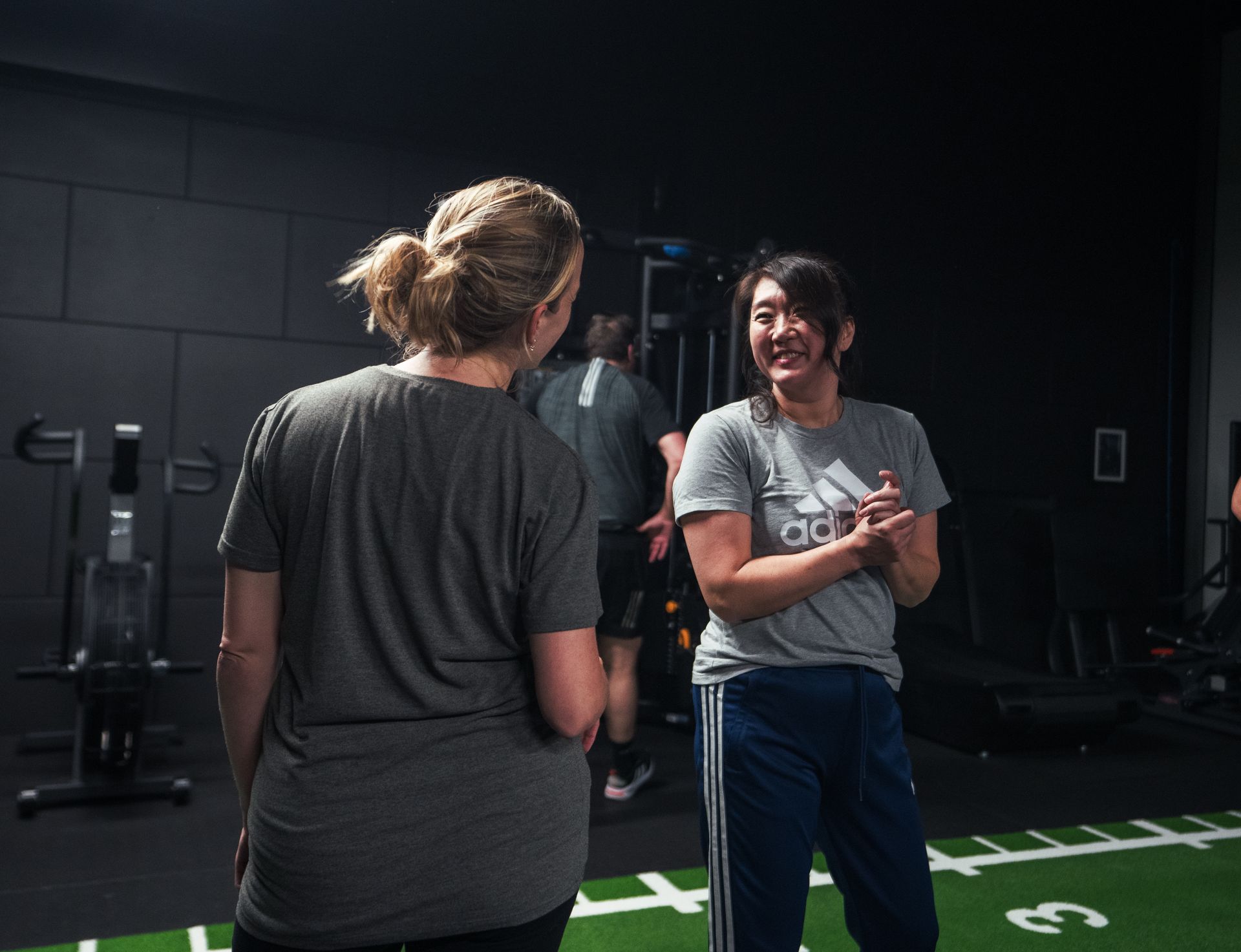 Twee vrouwen praten in een sportschool; één gebaart met haar handen. Op de achtergrond staan andere 