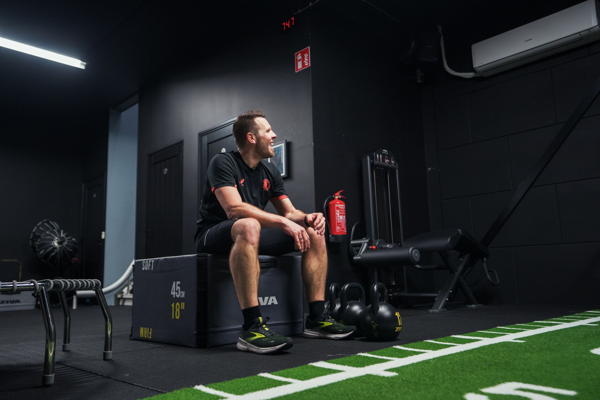Man in sportkleding die op een box in de sportschool zit, glimlachend, met kettlebells in de buurt.
