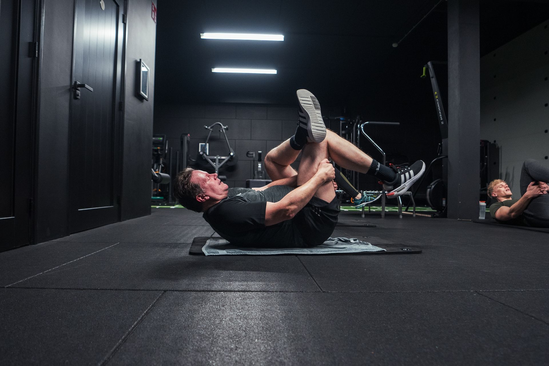 Een man strekt zich uit op een mat in een sportschool, waarbij hij zijn knie tegen zijn borst houdt. Op de achtergrond is nog iemand anders aan het rekken.