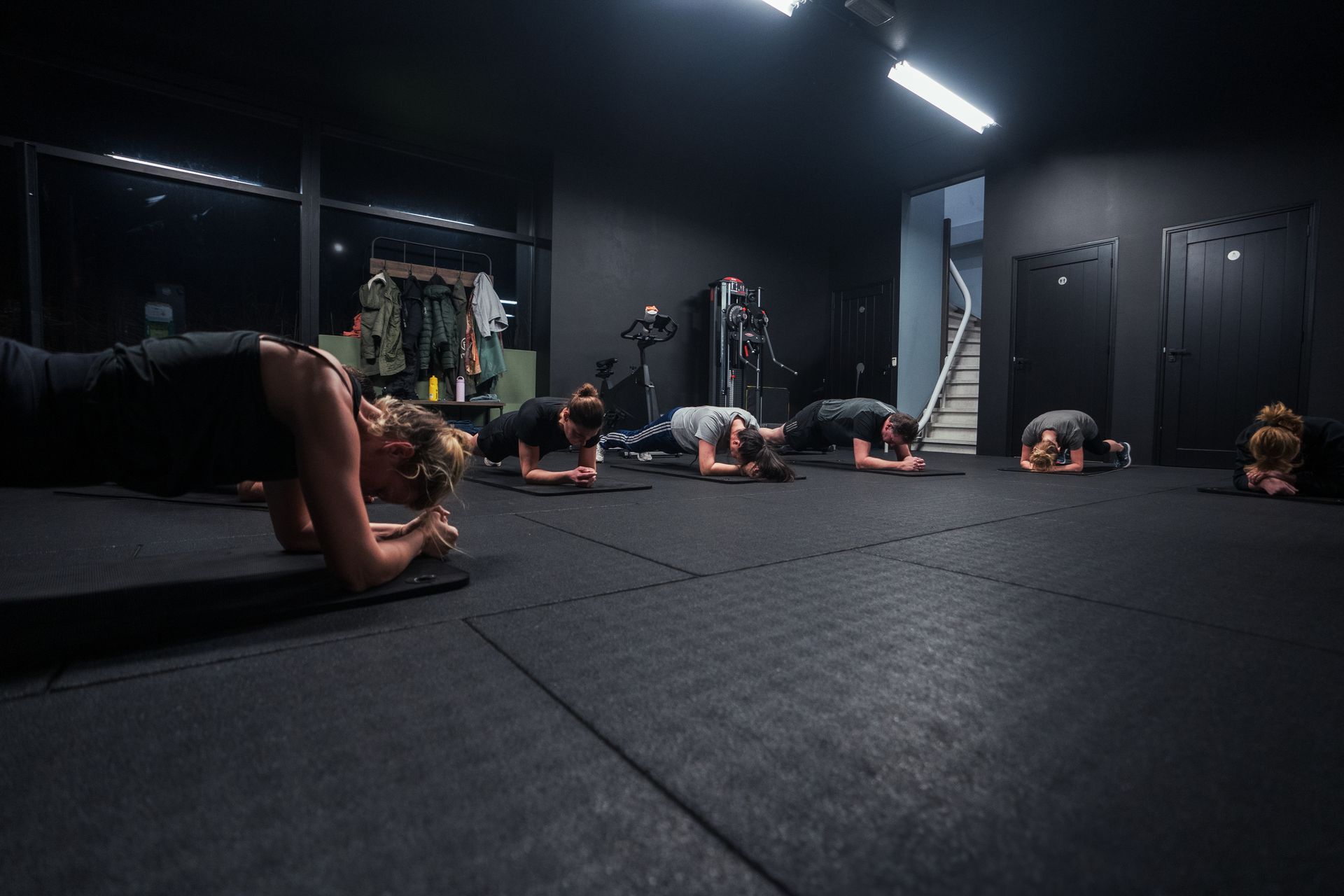 Mensen in een fitnessles doen plankoefeningen op een zwarte vloer in een donkere studio.