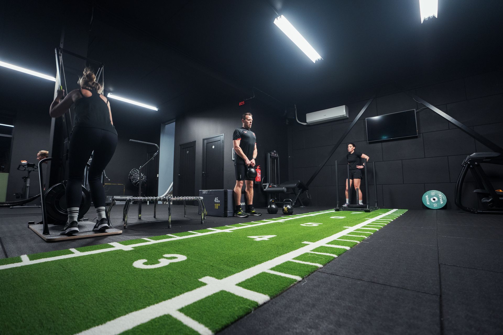 Mensen trainen in een donkere sportschool. De een gebruikt een apparaat, de ander staat op een kunstgrasveld met gewichten en een stang.