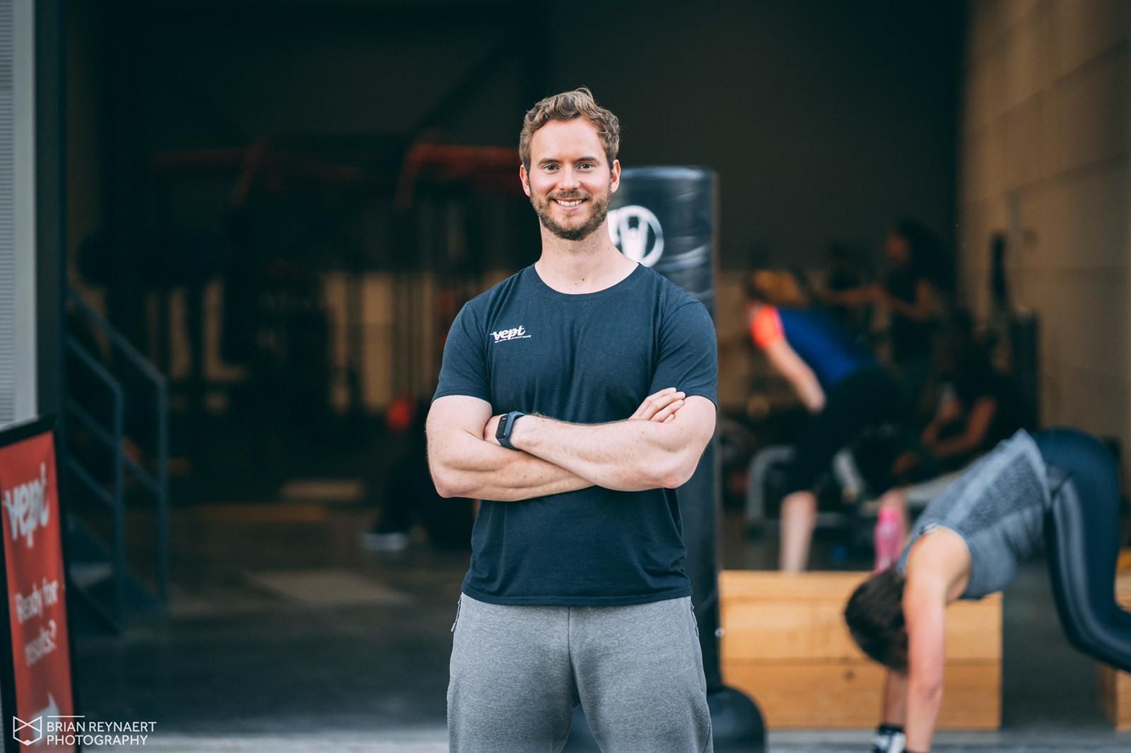 Een man met gekruiste armen in een sportschool, lachend. Op de achtergrond zijn mensen aan het trainen.