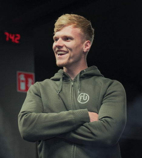Blonde man in een groene hoodie lacht met gekruiste armen voor een donkere achtergrond.