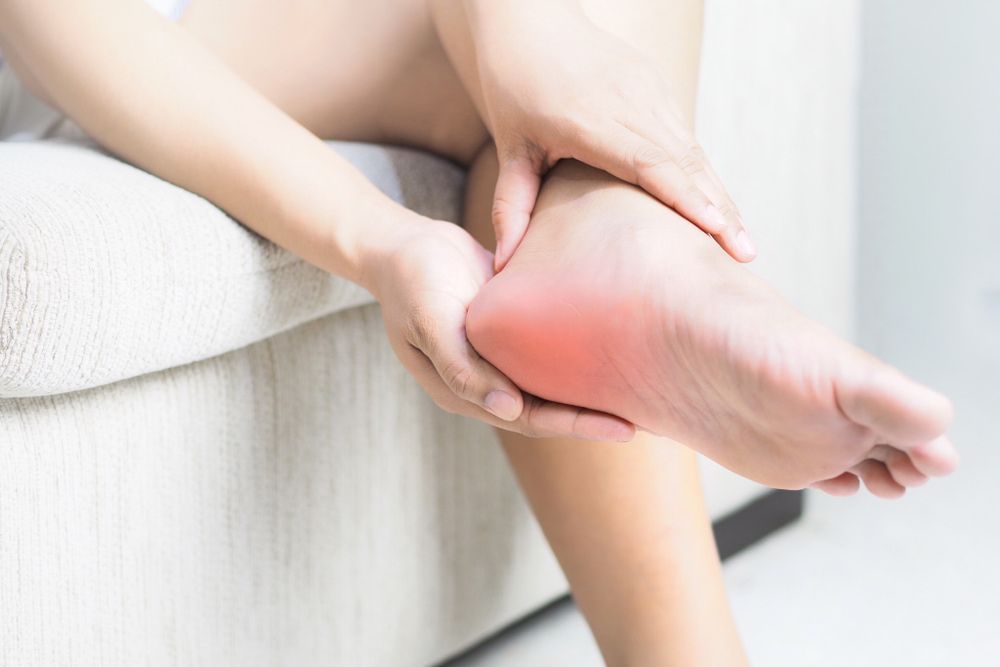 A Woman Is Sitting on A Couch Holding Her Foot in Pain — Swan Podiatry and Orthotics in Coconut Grove, NT