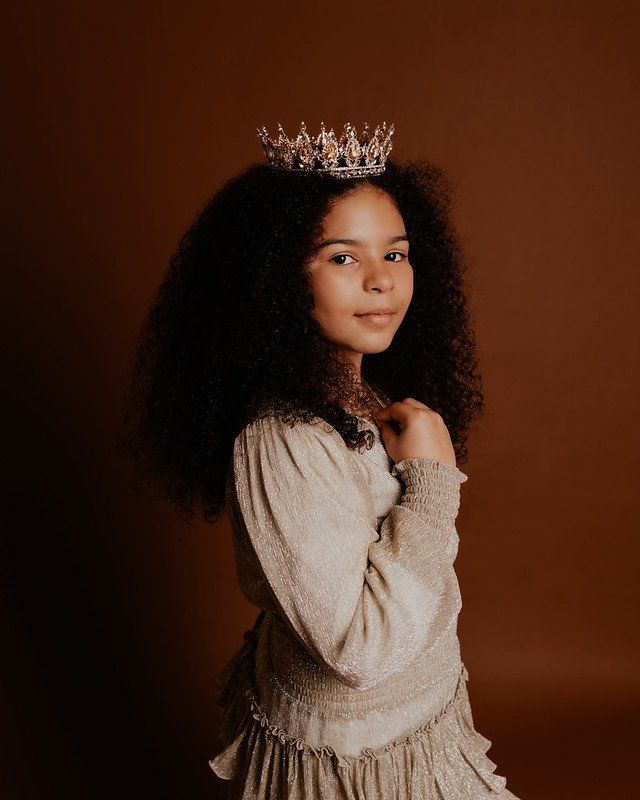 A young girl wearing a crown and a dress is standing in front of a brown background.