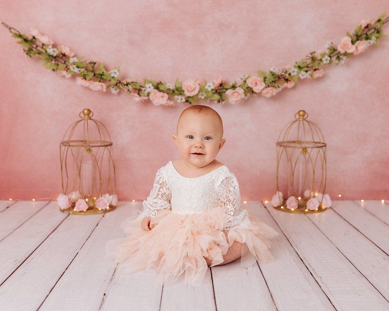 A baby in a pink dress is sitting on a wooden floor.