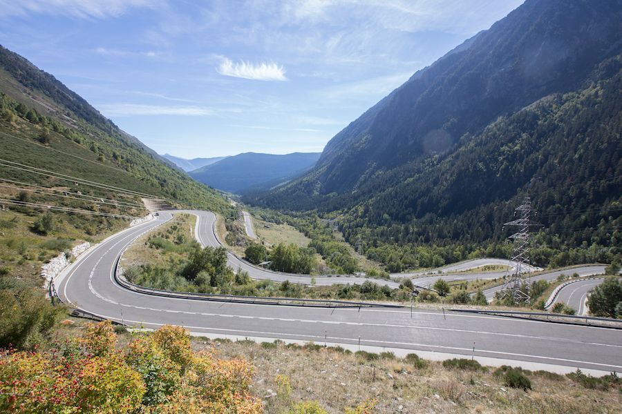 Winding mountain road through a green valley with steep rocky slopes and distant peaks under a blue sky