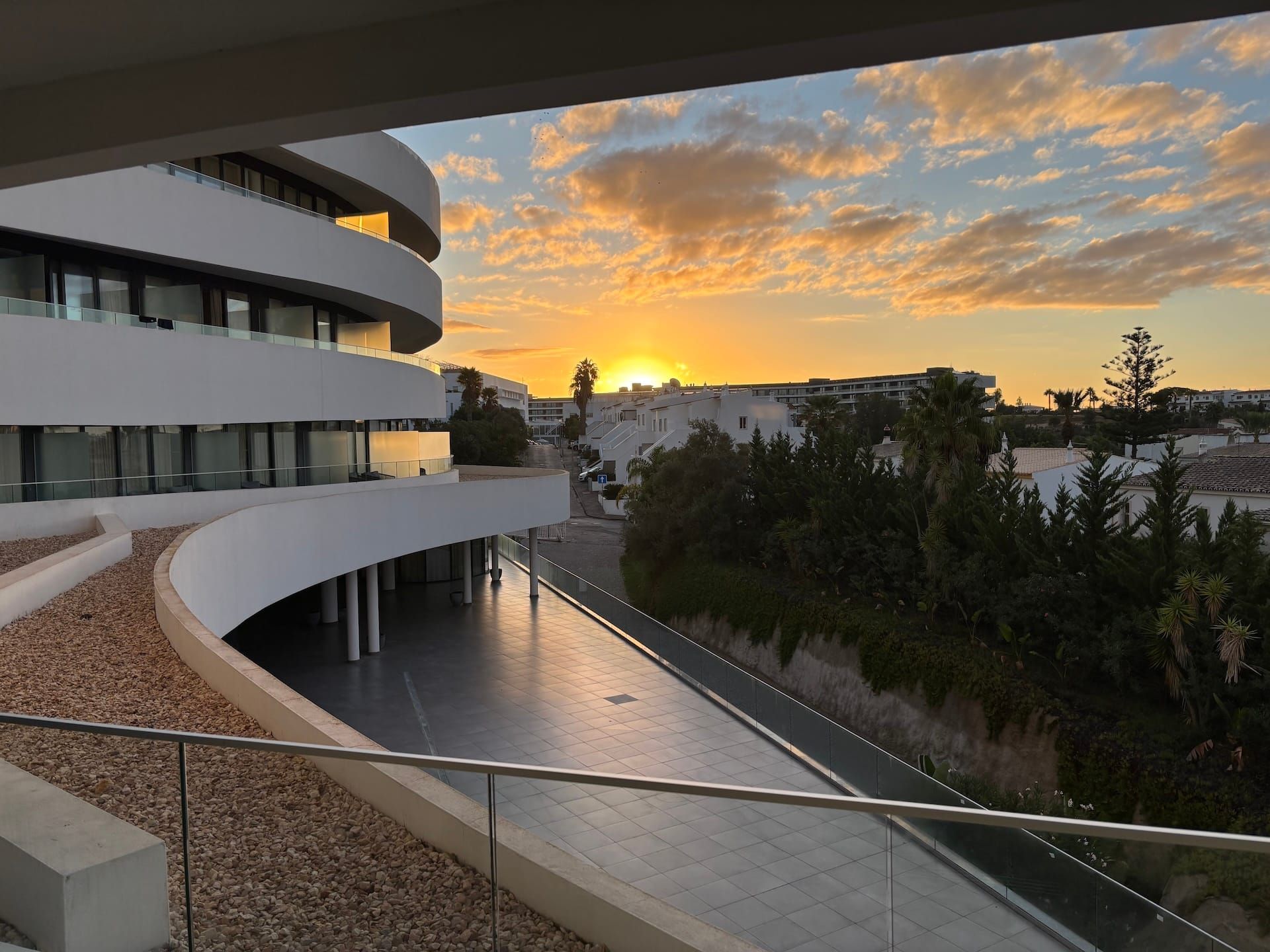 Curved modern building at sunset beside water, with glowing sky and trees in the background