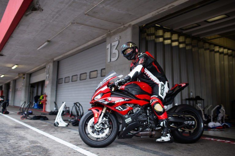 Motorcyclist on a red racing bike in a pit lane garage, with other bikes and gear nearby.