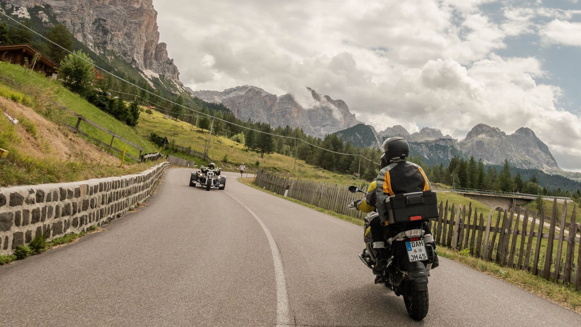 Motorcyclist riding on a winding mountain road with jagged peaks and cloudy sky