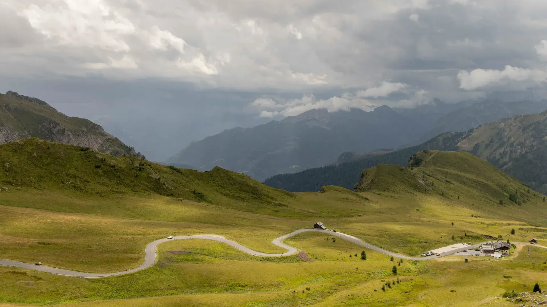 Winding mountain road through green hills under cloudy skies, with distant peaks and tiny hikers