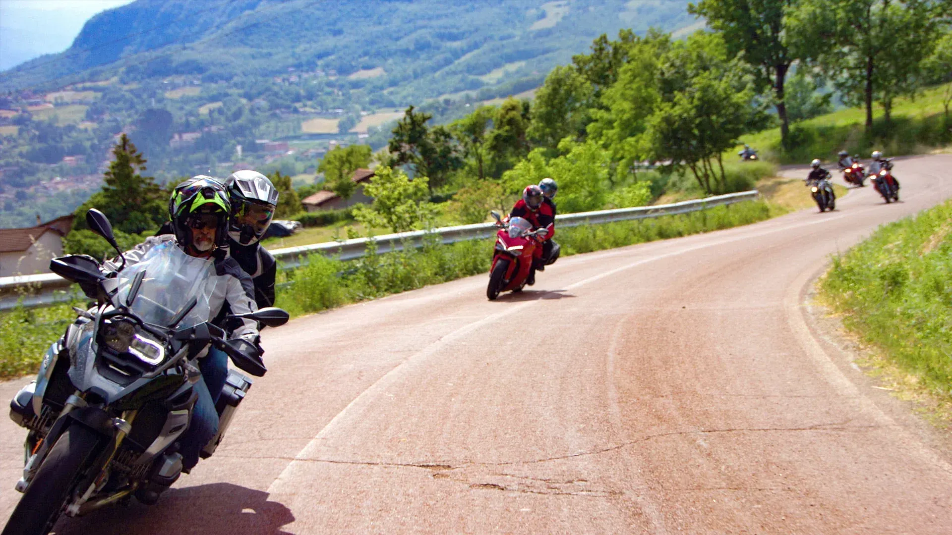 Motorcyclists lean into a curve on a winding mountain road with green hills in the background.