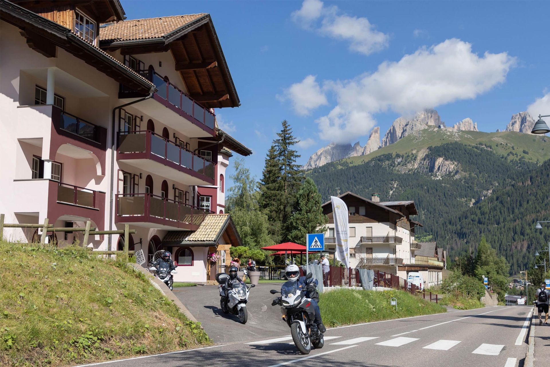 Motorcyclists on a mountain village road beside chalet-style buildings under a blue sky