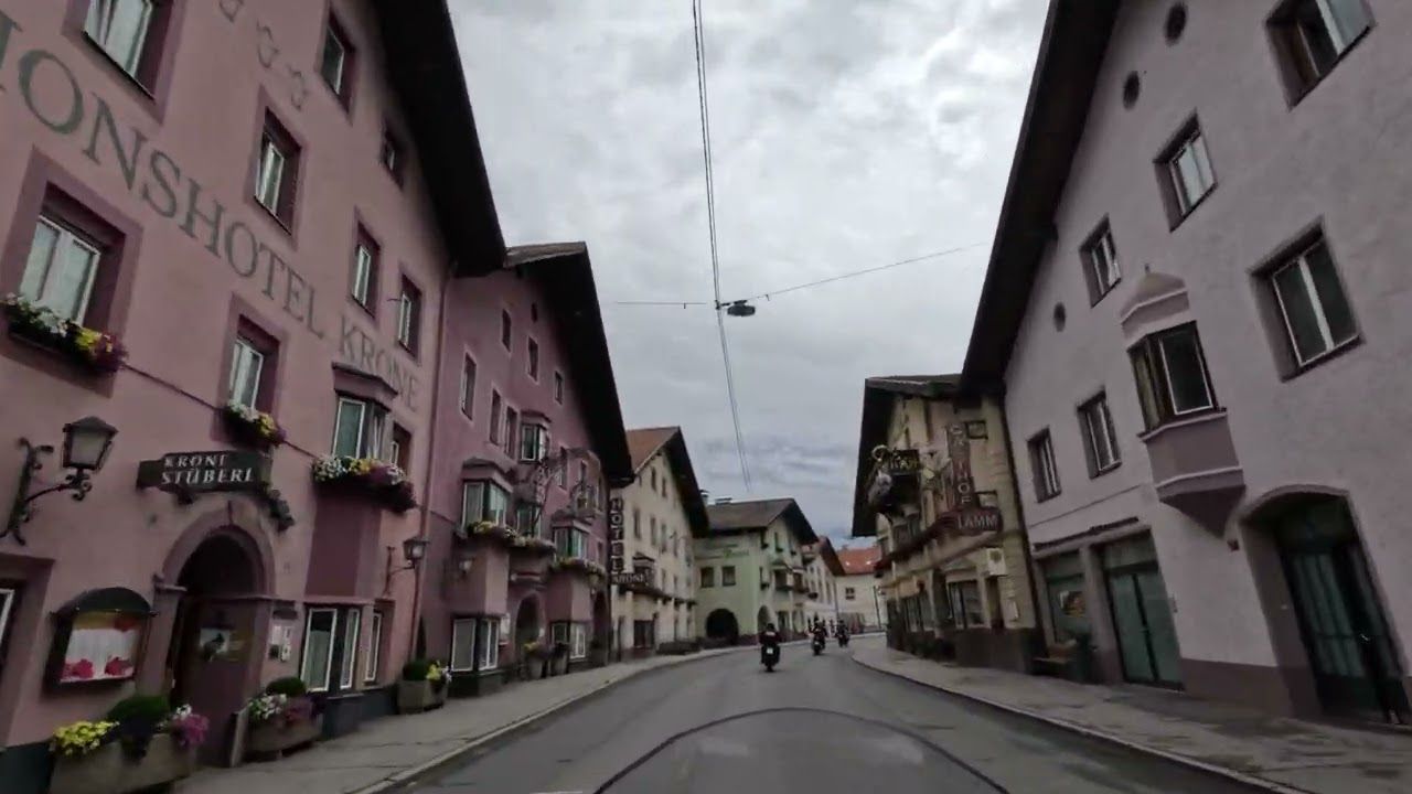 Curved cobblestone street lined with pastel buildings under a cloudy sky, with a few pedestrians ahead