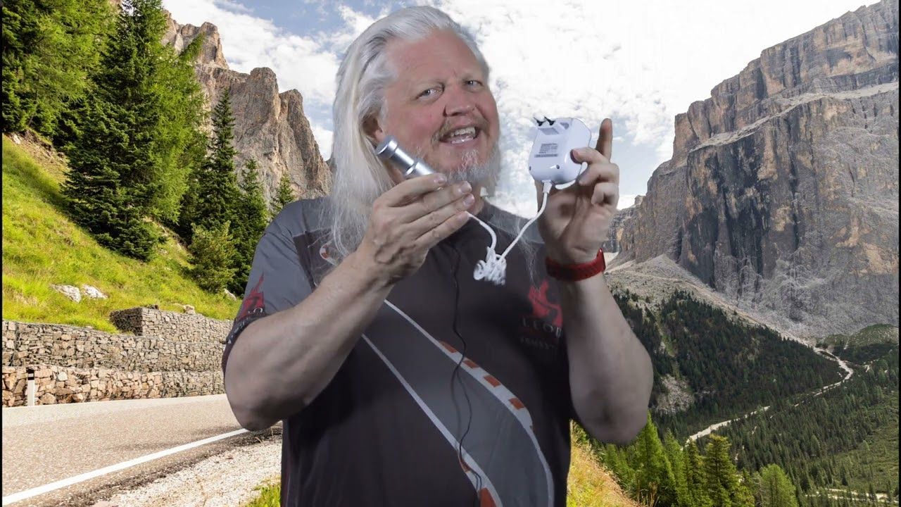 Man holding a cup and phone, smiling on a mountain road with cliffs and green hills behind him