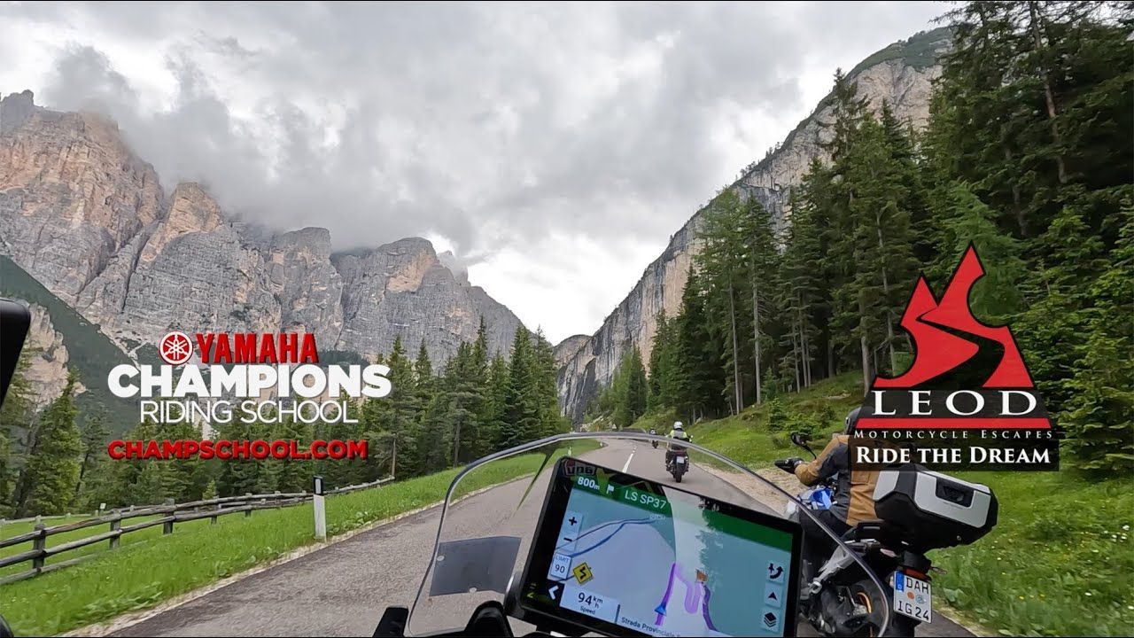 Motorcycle dash view on a mountain road with cliffs, trees, and cloudy sky; Yamaha Champions and LED logos visible