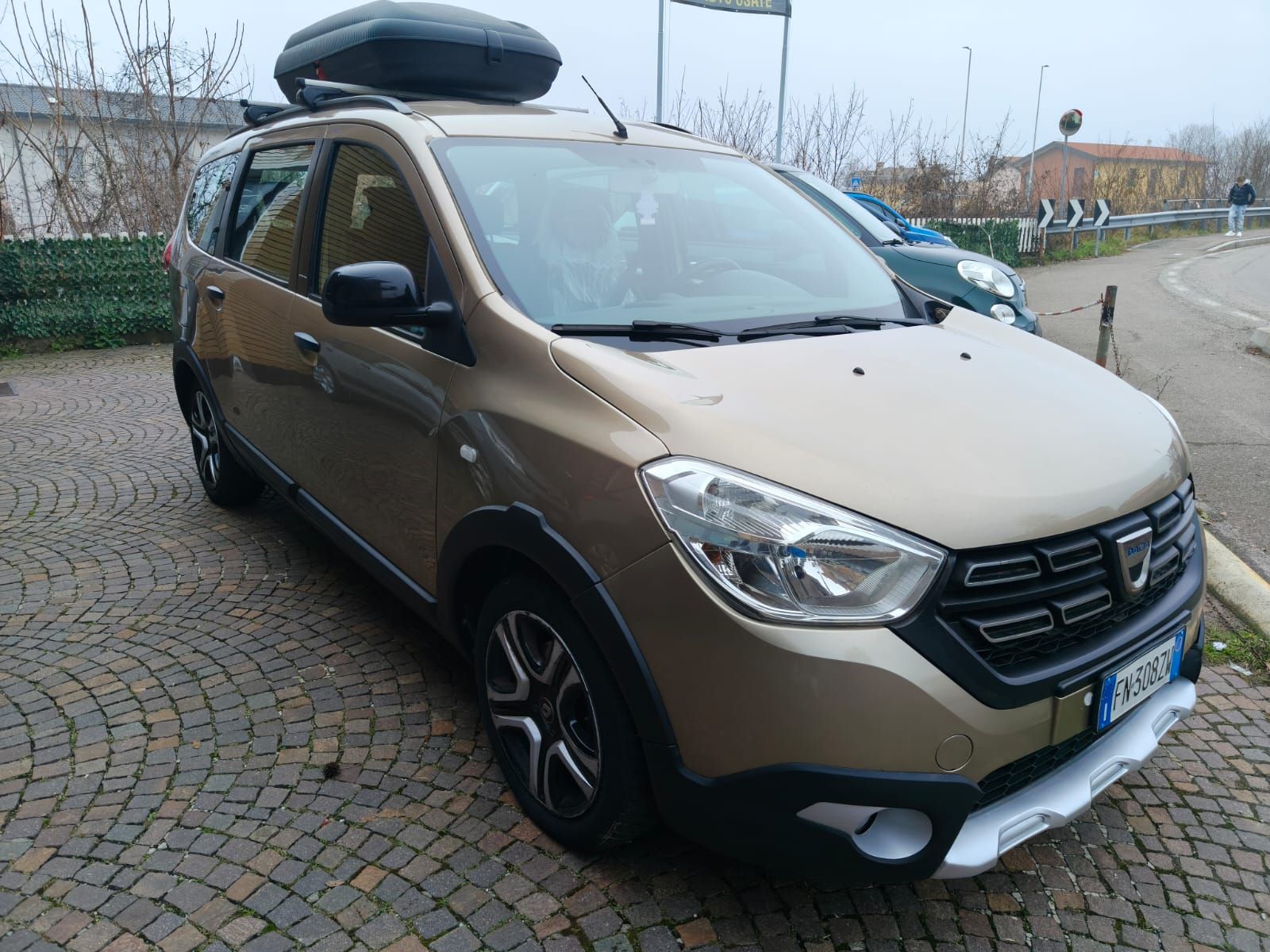 Dacia Lodgy color oro con box sul tetto parcheggiata su una superficie di ciottoli.