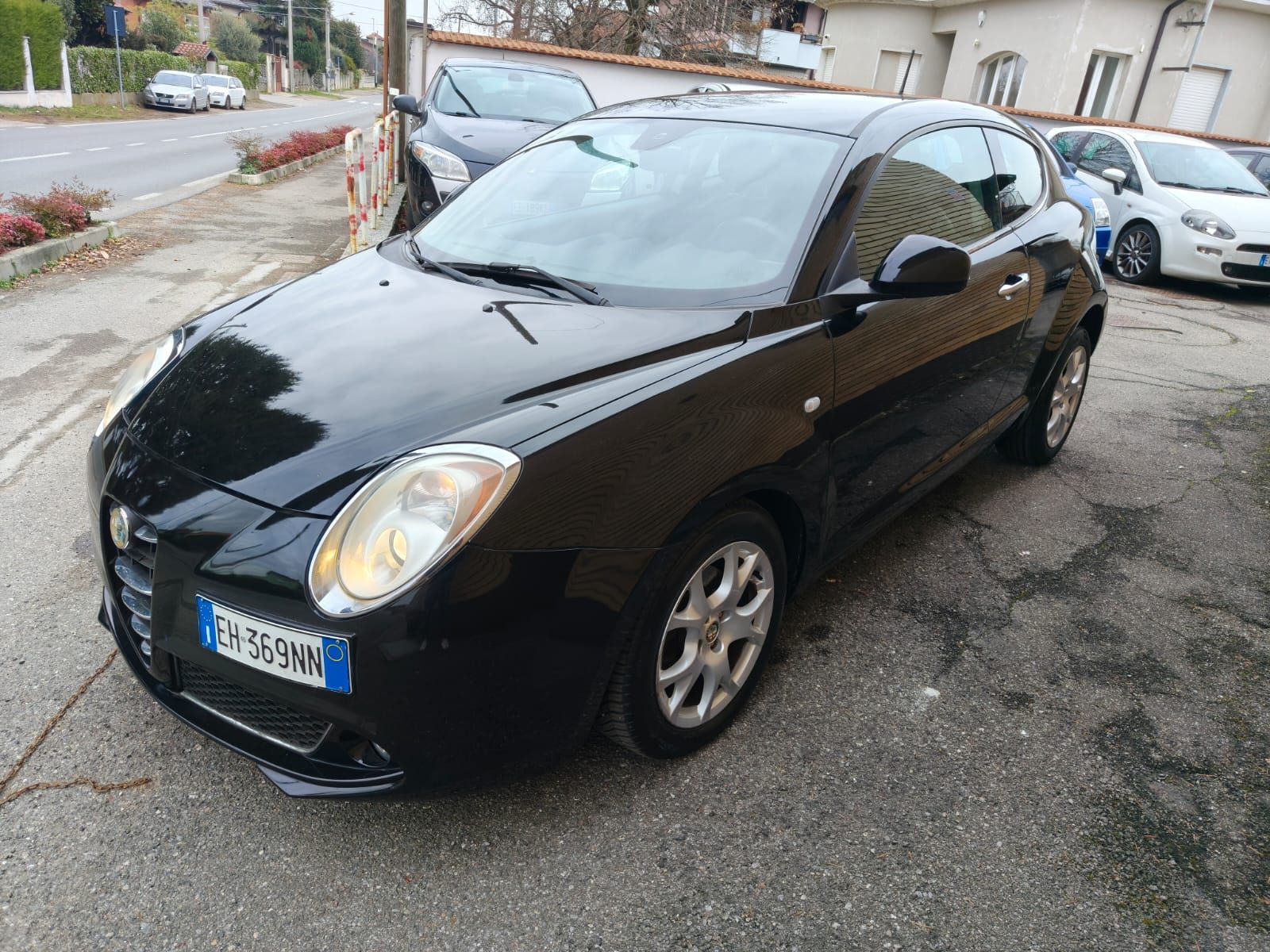 Un'Alfa Romeo MiTo nera parcheggiata su un terreno asfaltato accanto a una strada, con altre auto sullo sfondo.