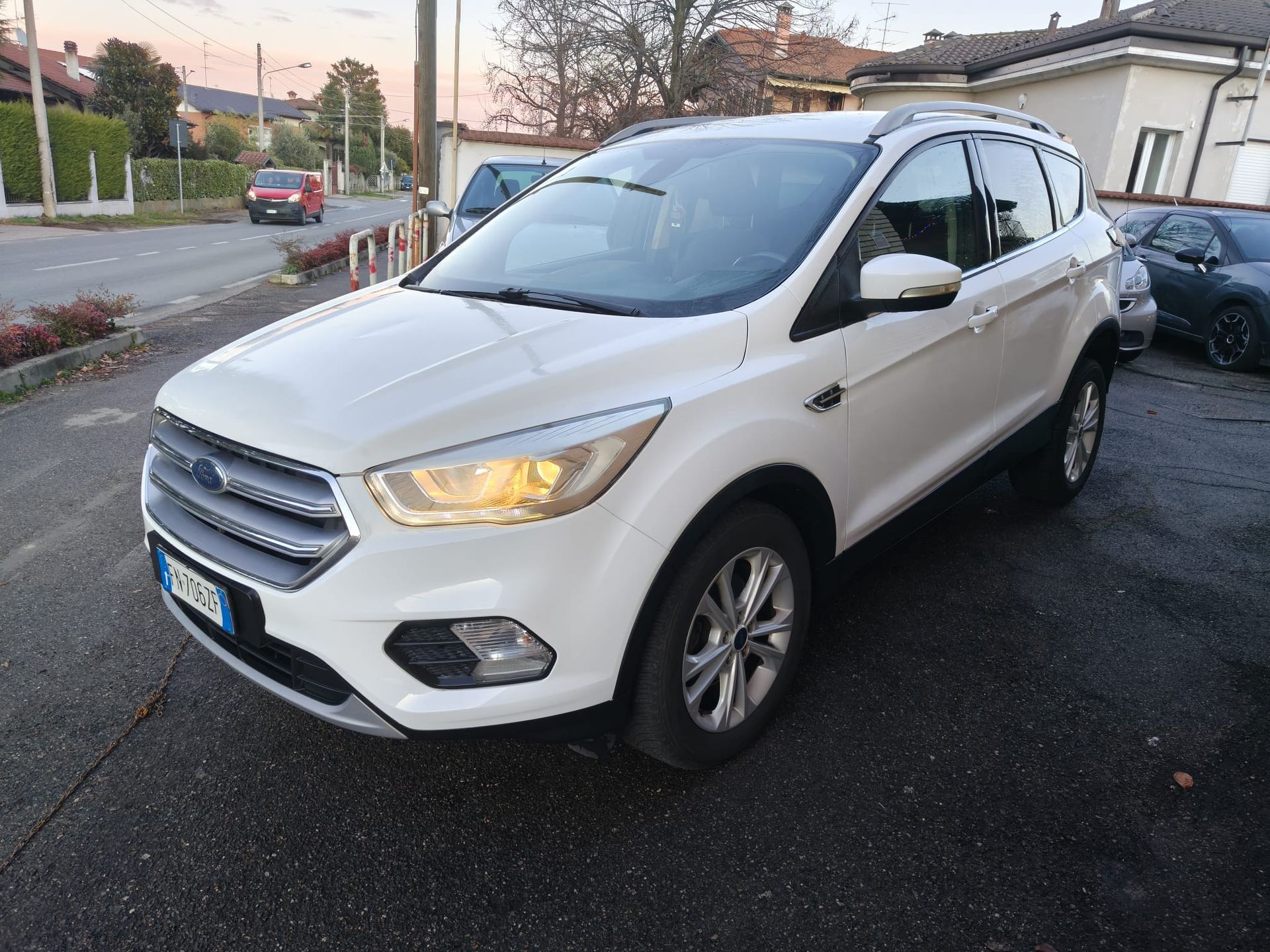 Un SUV Ford Kuga bianco parcheggiato su un terreno asfaltato insieme ad altri veicoli, sotto un cielo coperto.