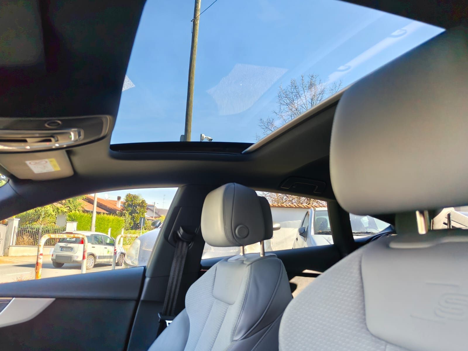 Vista interna di un'auto con tettuccio apribile parzialmente aperto, che mostra un cielo azzurro e sedili chiari.