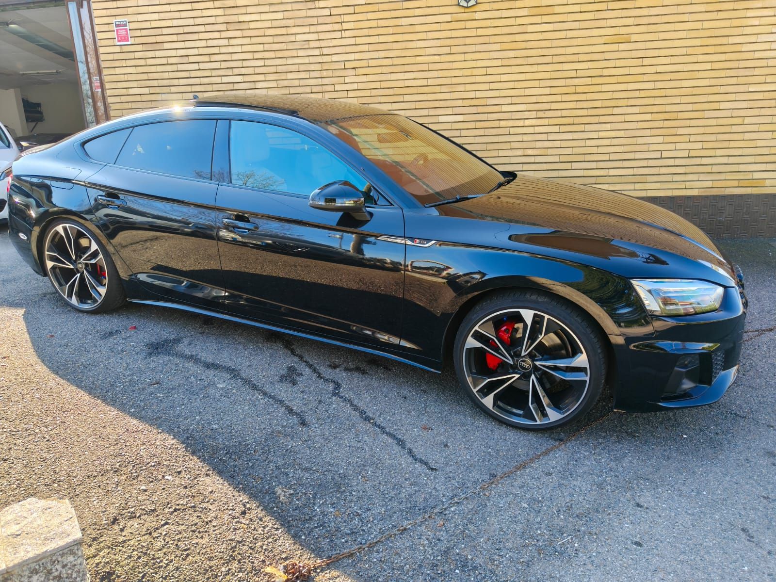 Berlina Audi nera parcheggiata sul marciapiede, vetri oscurati, pinze dei freni rosse, luce del sole.