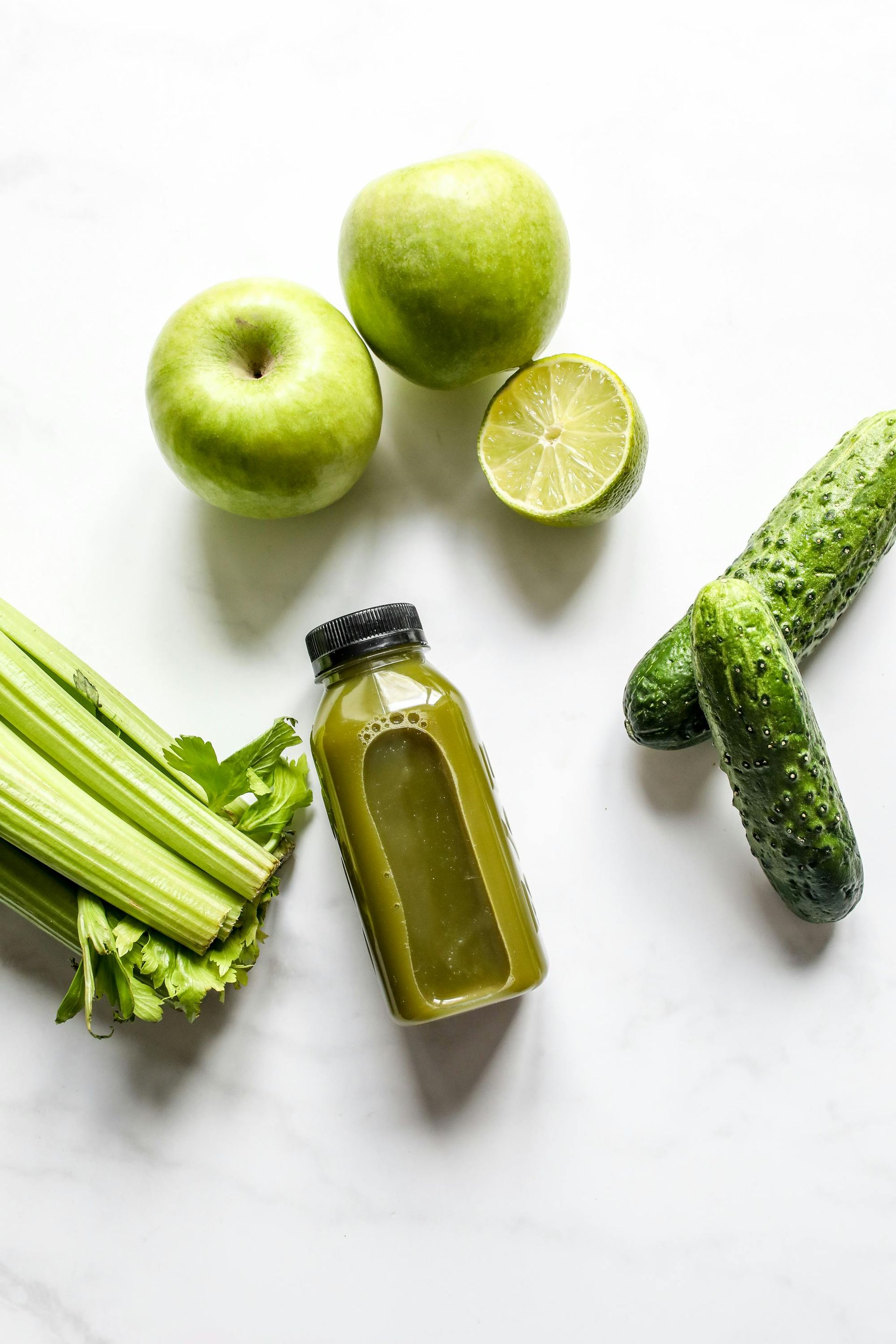 A bottle of green juice surrounded by celery , apples , limes and cucumbers on a table.