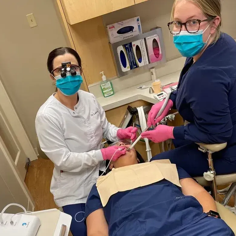 Two women wearing masks are working on a patient 's teeth in a dental office.