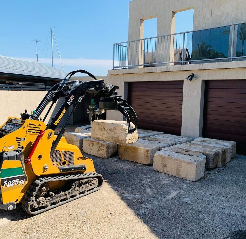 A Yellow Skid-steer Loader is Lifting a Large Stone Block in Front of a Garage With Wooden Doors