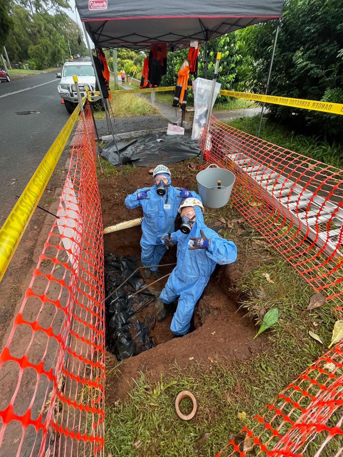 Two workers in blue hazardous material suits and respirators pose inside a dug-out trench surrounded by orange safety mesh — Lifestyle Demolition In Mullumbimby, NSW 