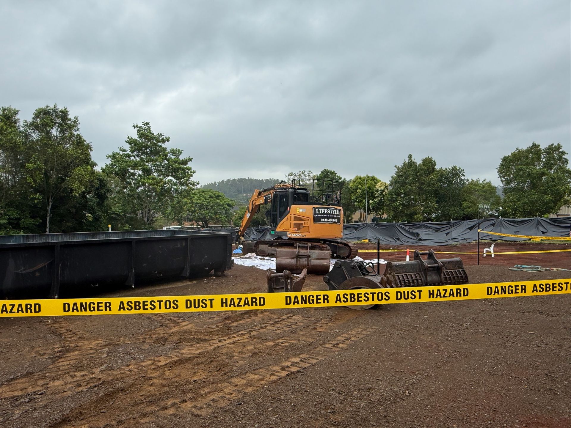 An orange excavator parked on a dirt lot behind yellow danger asbestos warning tape with black plastic sheeting nearby — Lifestyle Demolition in Kingscliff, NSW