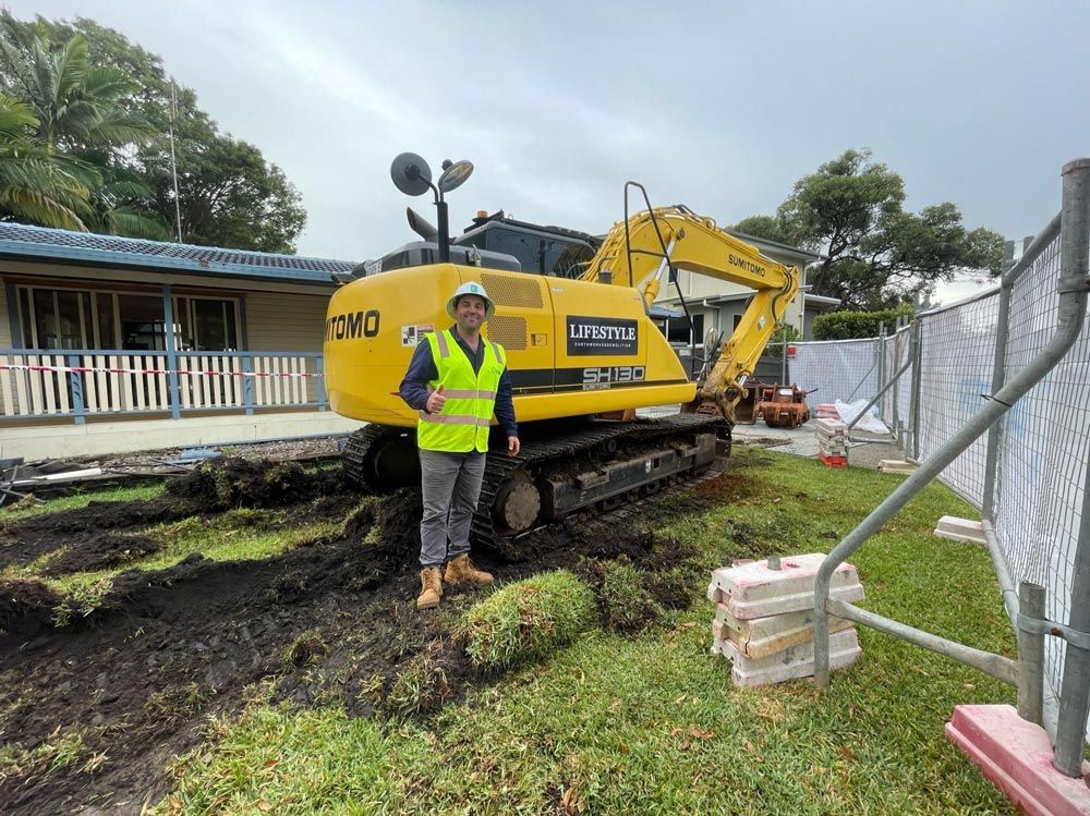A Man Is Driving a Small Excavator in Front of A House — Lifestyle Demolition In Byron Bay, NSW