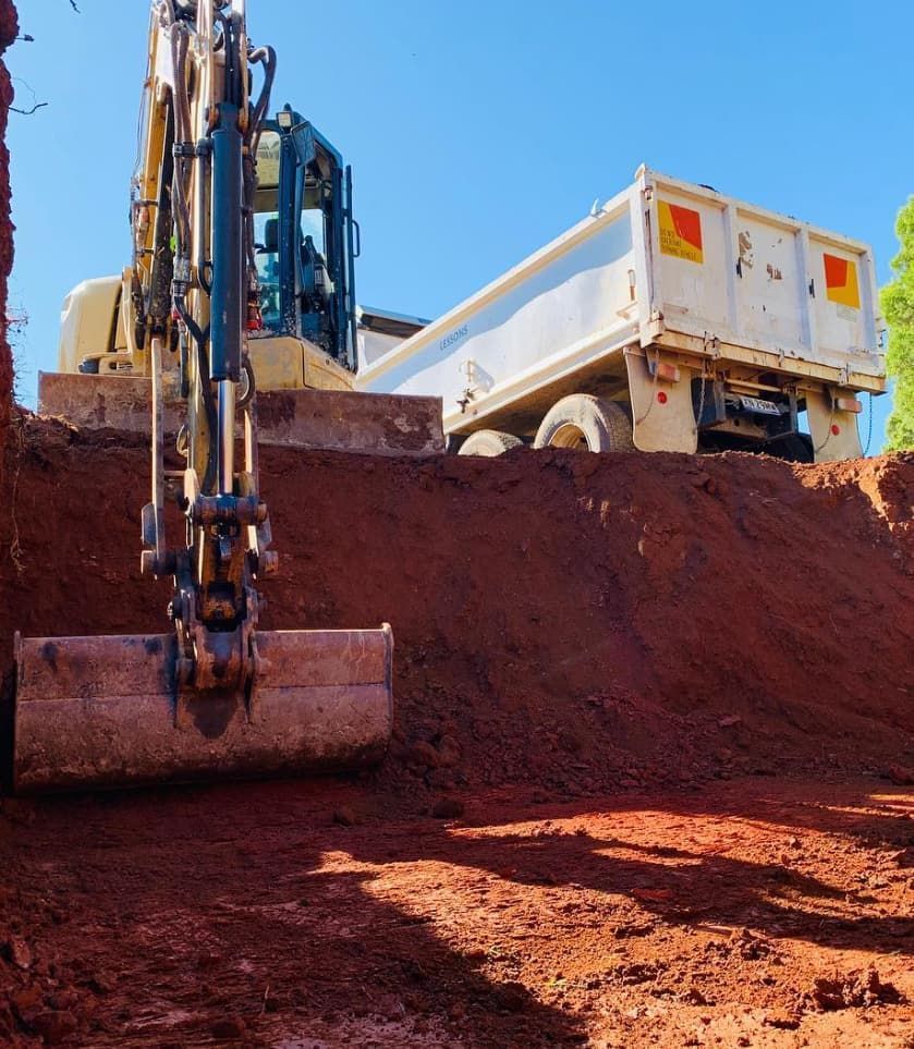 An Excavator Loads Reddish Soil Into A White Dump Truck On A Sunny Day — Lifestyle Demolition In Ballina, NSW