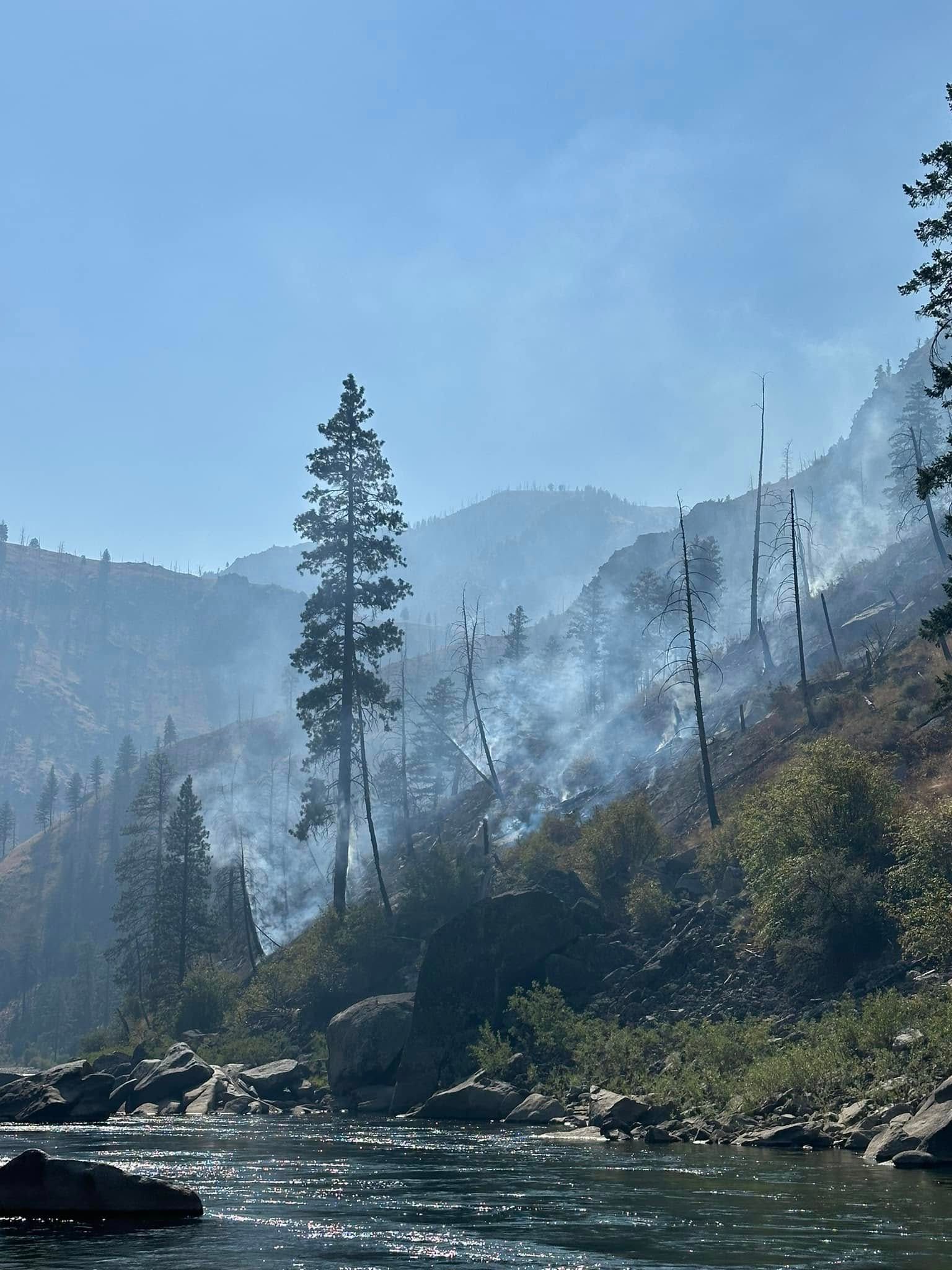 A river surrounded by trees and mountains with smoke coming out of the trees.